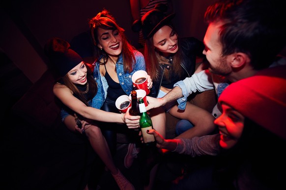 High angle of several young people, girls and one man, getting drunk at club party, sitting close together in dark red lit room and cheering to their meeting