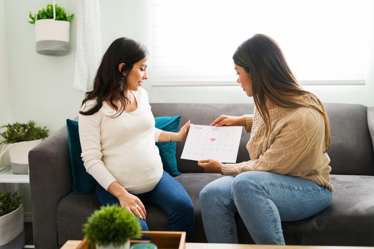 Beautiful pregnant woman and latin midwife holding a calendar. Doula and expectant mother talking about the due date while sitting on the couch