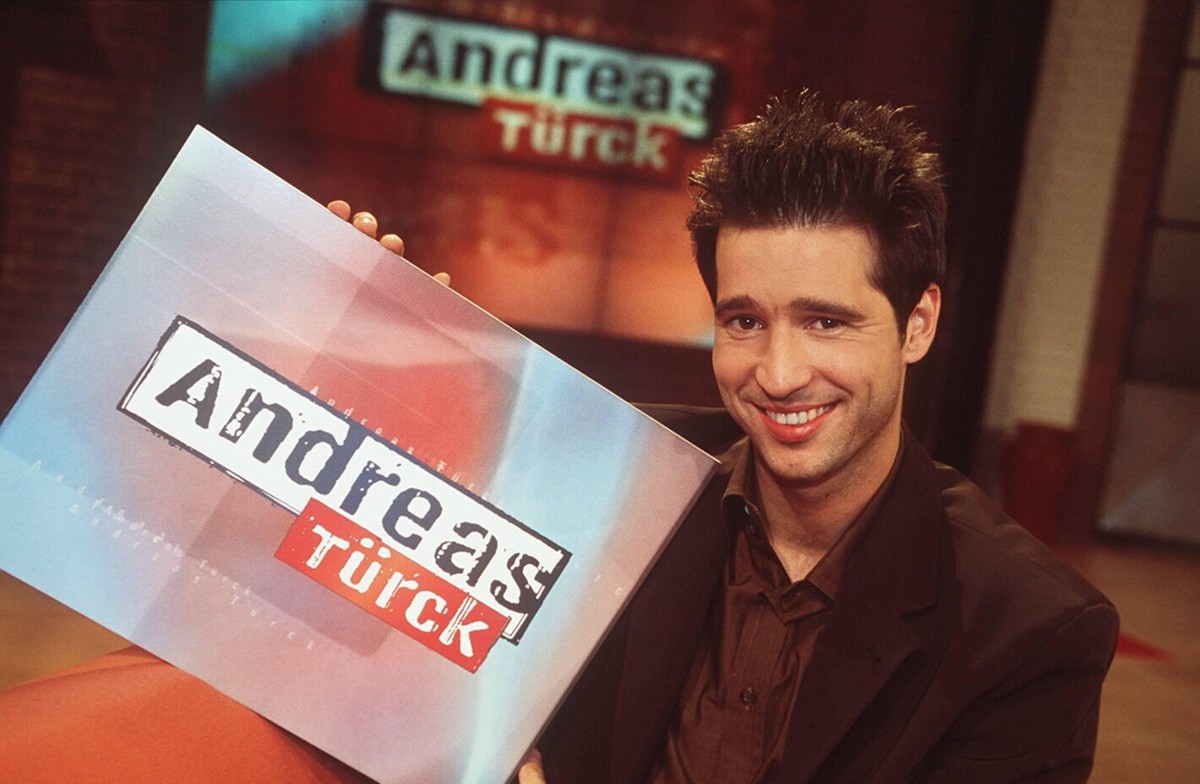 Andreas Türck in Andreas Türck (PRO SIEBEN) 01/98 her Mann TV Fernsehen Moderation dunkelhaarig leger Jackett Hemd braun Studio Dekoration Kulisse Schild haltend Logo halb quer lächelnd sitzend