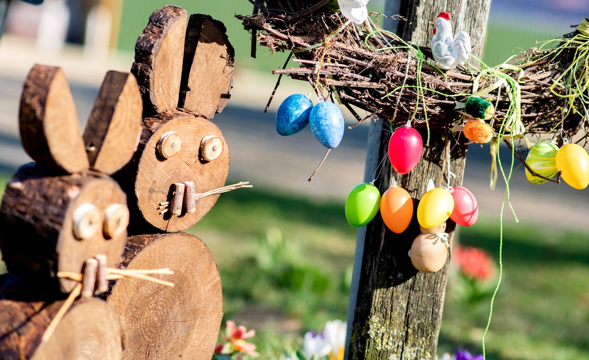 Zwei Osterhasen aus Holz stehen in einem Vorgarten im Ortskern, während bunte Ostereier an einem Strauch hängen.