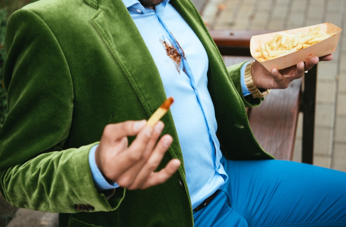 partial view of man in green velvet jacket with ketchup on shirt and french fries in hands sitting on bench, partial view of man in green velvet jacket with ketchup on shirt and french fries in hands ...