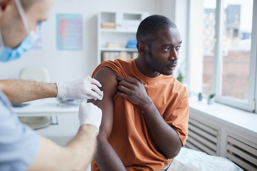 Portrait of adult African-American man looking away while getting covid vaccine in clinic or hospital, with male nurse injecting vaccine into shoulder