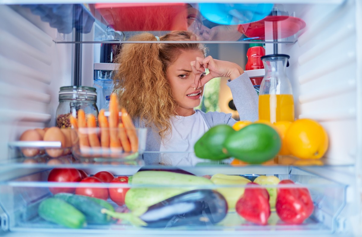 Woman standing in front of opened fridge and holding up to her nose because of bad smell. Picture taken from the inside of fridge full of groceries.