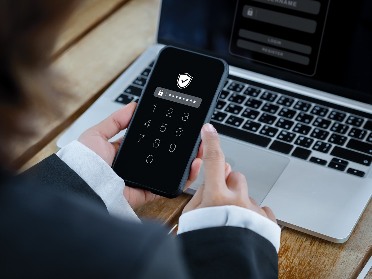 Two factor authentication or 2FA and cyber security concept. Shield icon and numbers on smartphone screen in hand, access password to laptop, identity verification with PIN verification code number. x ...