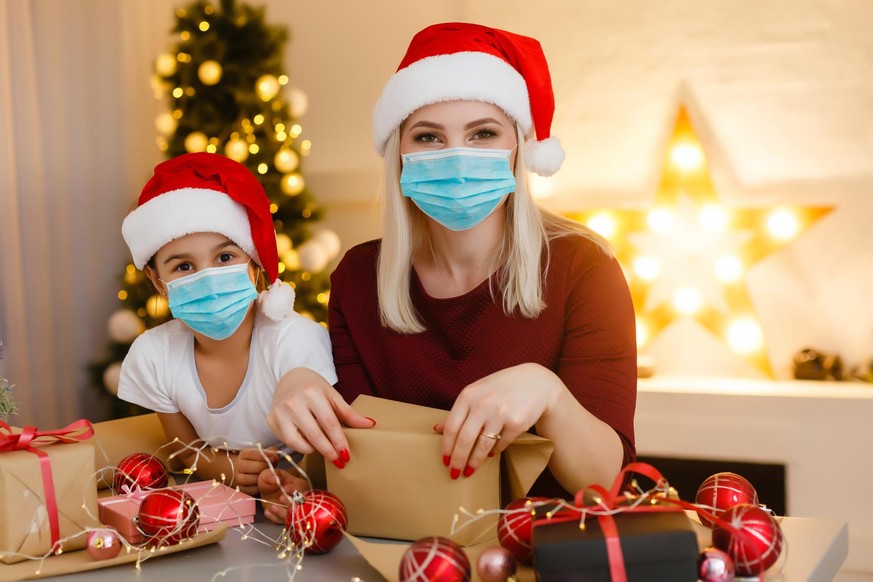 Beautiful mother and daughter in medical masks have fun at home near the Christmas tree in a white interior. Family happiness, holiday, joy, vacation, games with a woman. New Year's preparations. Cele