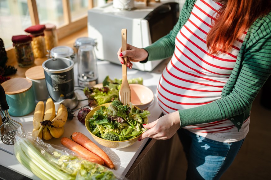 Happy eight month pregnant woman with red dyed hair making a healthy vegan salad in her modern kitchen