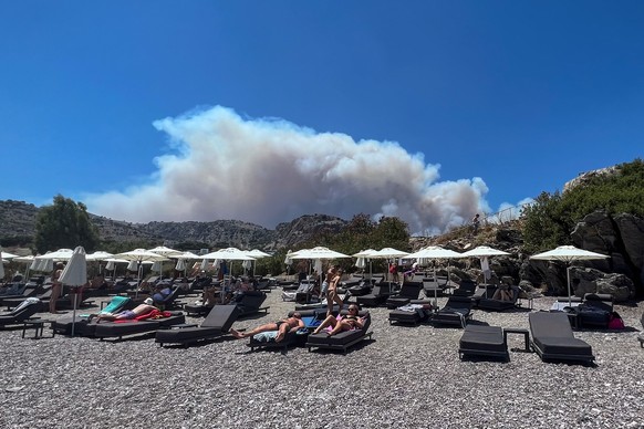 22.07.2023, Griechenland, Lindos: Am Viglika Beach auf der griechischen Insel Lindos aus, wo sich Menschen auf Sonnenliegen am Strand aufhalten, kann man aufsteigenden Rauch von Waldbränden auf der Fe ...