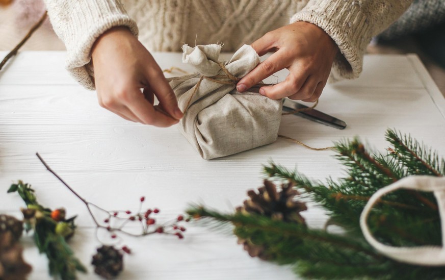 Female hands wrapping stylish christmas gift in linen fabric on white rustic table with green branch, pine cones, scissors and twine. Woman in cozy sweater preparing plastic free christmas present