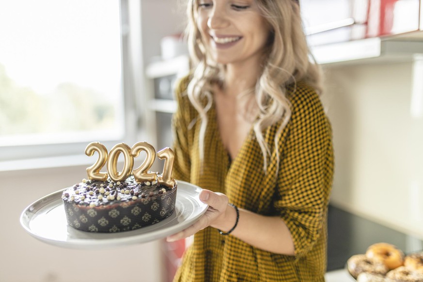 Young woman making and decorating chocolate cake with lit candles shaped as numbers 2021 for New Years party