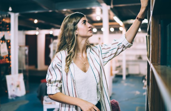 Young Caucasian woman taking book from high shelf in library searching literature for self education in college, smart female librarian putting literature in order during work in bookstore