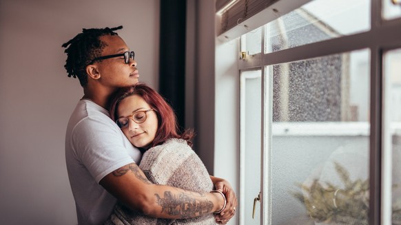 Interracial couple hugging each other while standing near window. African man embracing his caucasian girlfriend at home.