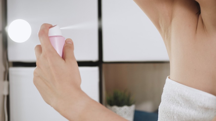 Girl uses deodorant. Young attractive woman in white towel in bathroom with her left hand sprays deodorant on her right armpit. Hygiene, skin care, freshness concept. Close-up view