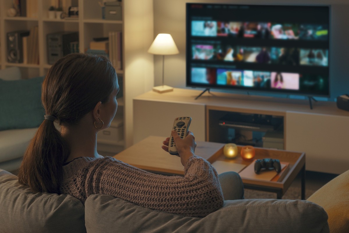 Woman relaxing on the couch, she is using the remote control and choosing a TV show or movie on the television menu