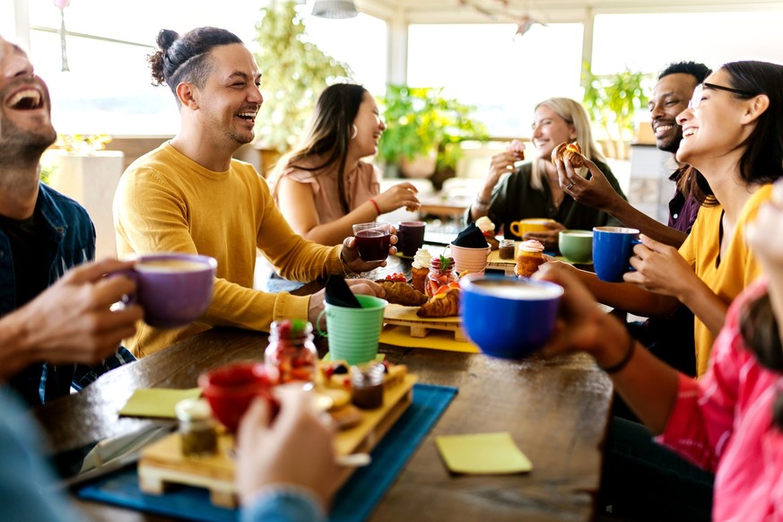 Smiling group of diverse friends having breakfast and talking at coffee bar restaurant - Happy millennial multiracial people laughing and laughing together while drinking cappuccino