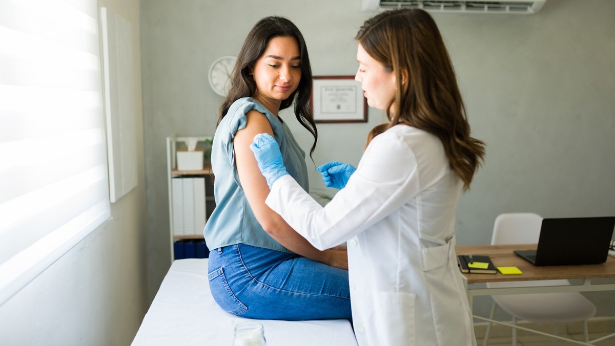 Young Hispanic woman is getting vaccinated by a professional medical worker wearing gloves in a doctor&#039;s office