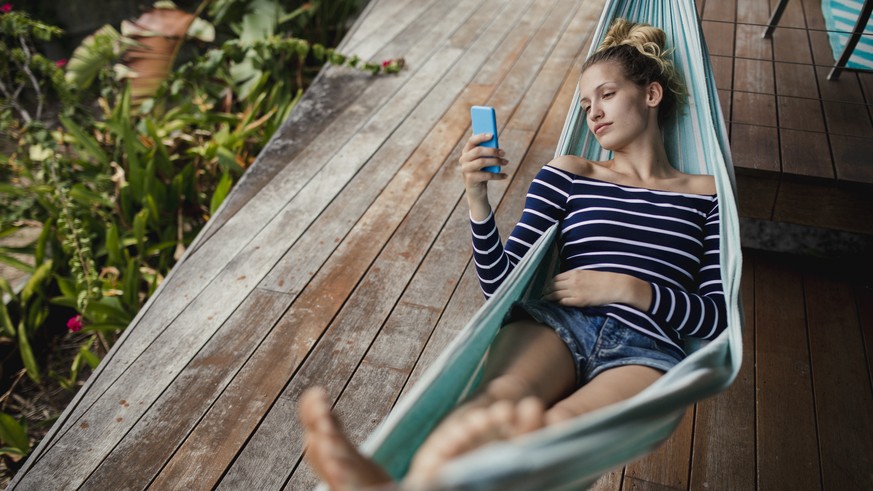 Young woman lying down on a hammock, using her smartphone and relaxing.