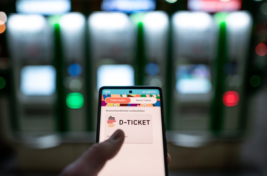 ARCHIV - 03.04.2023, ---: Ein Mann hält ein Mobiltelefon, auf dem das «D-Ticket» gekauft werden kann, vor Ticketautomaten am Hauptbahnhof in der Hand. (zu dpa: «Bayern: Bund muss Zusagen bei Deutschla ...