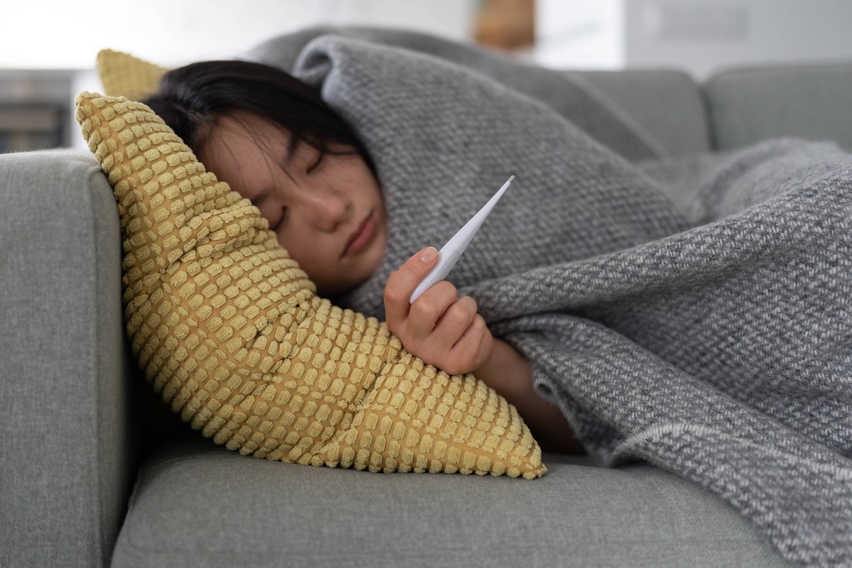 Sick Asian girl lying on sofa wrapped in blanket hold thermometer after infection, covid-19 outbreak Sick Asian girl lying on sofa, wrapped in blanket and holds thermometer to measure body temperature ...