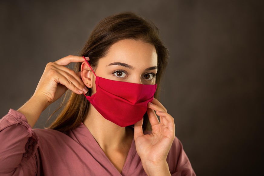 Woman attaching red cloth face mask on ear with fingers with dark background. Young girl putting on respiratory protection during epidemic. Concept of stopping spreading of coronavirus.