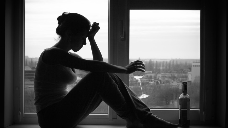 Sad young woman sitting on the window, drinks wine. Alcoholism problem. Black and white photography.
