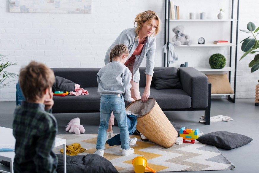 tired mother putting toys in basket while naughty children playing at home