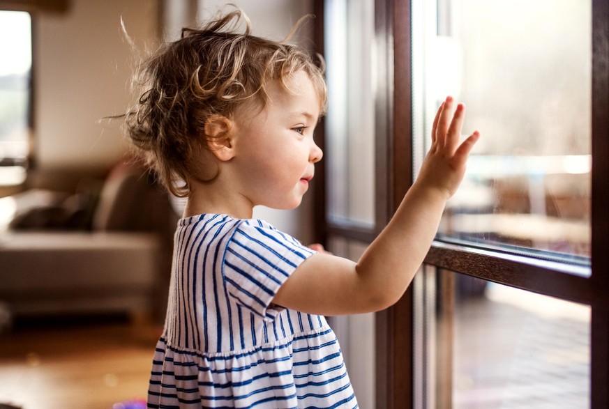 A happy toddler girl standing by window indoors at home, looking out.