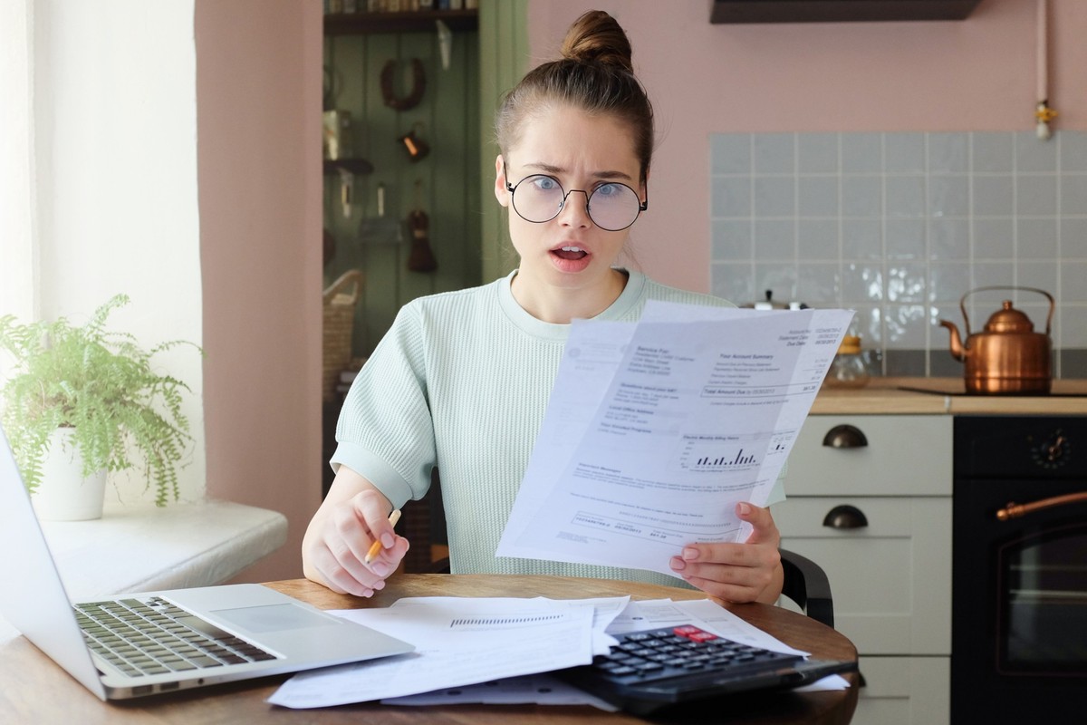 Amazed young woman sitting at kitchen table, holding utility bills in hand, looking at parers with opened mouth, puzzled expression, surprised by high taxes. Financial problems concept