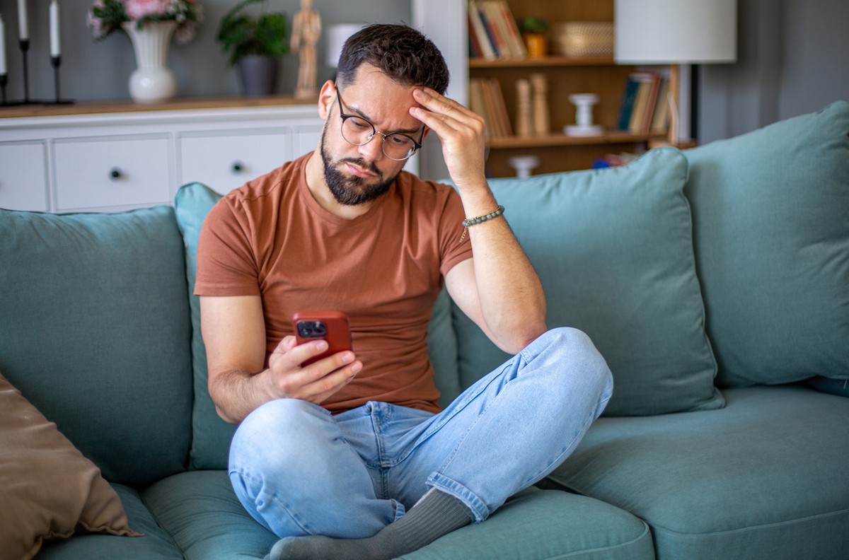 Worried man sitting on sofa at home reading bad news on his mobile phone