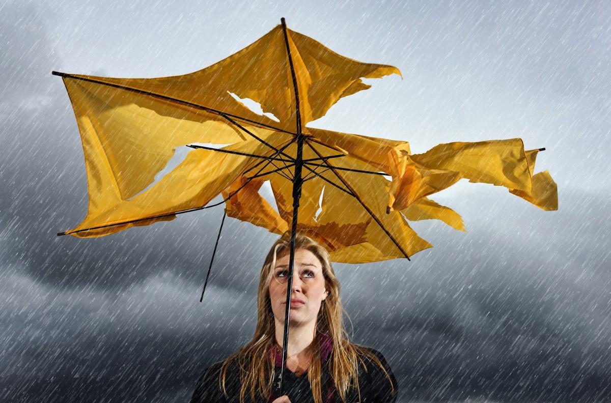 A beautiful young blonde woman sheltering under a torn and tattered yellow umbrella looks up miserably as she waits for a thunderstorm to pass.
