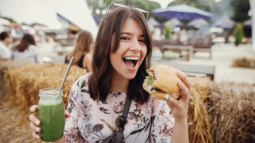 Stylish hipster girl in sunglasses holding delicious vegan burger and smoothie in glass jar in hands at street food festival. Happy boho woman holding burger and drink in summer street