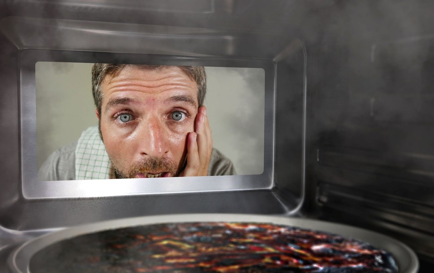 Junge chaotische und lustige Dummy-Mann in der Küche mit Blick durch Mikrowelle oder Ofen Pizza Brennen überkocht macht ein Durcheinander von Hauskoch in häusliche Katastrophe und Lifestyle-Konzept