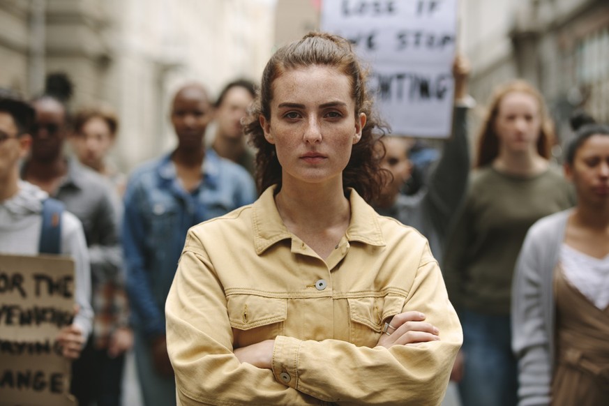 Young woman standing with arms crossed standing in a rally. Woman protesting with group of activists outdoors on road.