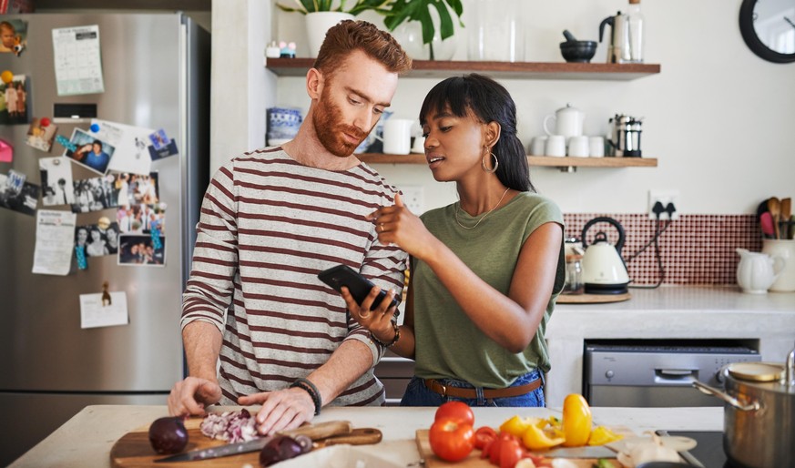 Shot of a young woman using her cellphone while cooking with her boyfriend at home