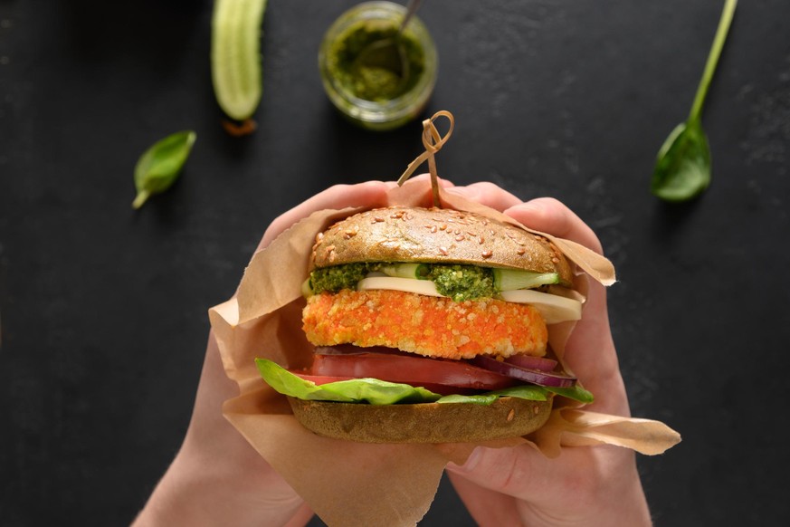 Trendy Vegan Burger of green spirulina buns, vegetables, vegan cheese, pesto carrot meatball in hands. Close up. Horizontal orientation.