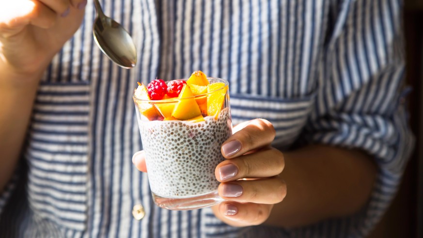 Chia pudding on coconut milk with raspberries and peach in a glass jar in female hands with a spoon. Raw vegan healthy food concept