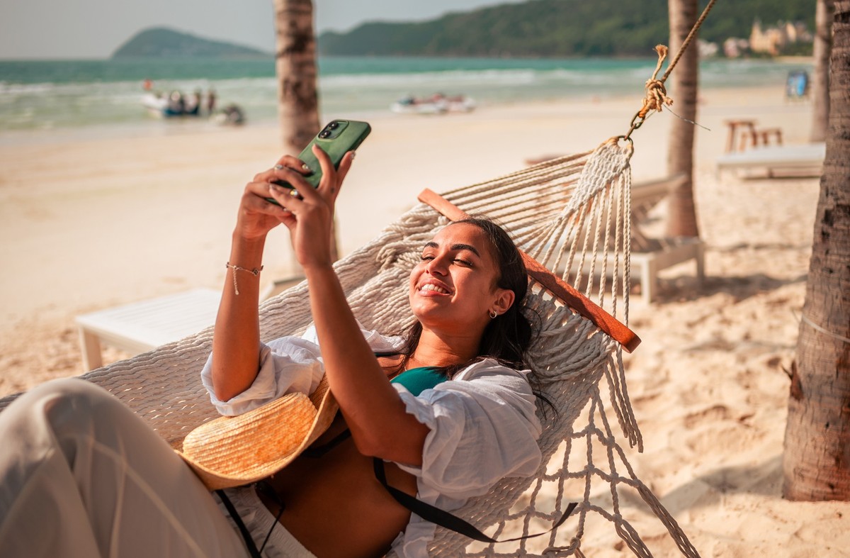A delighted young Hispanic woman lounges in a hammock by the ocean, taking a selfie with her smartphone while wearing a chic sun hat.