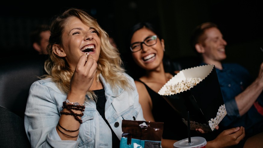 Young woman with friends watching movie in cinema and laughing. Group of people in theater with popcorns and drinks.