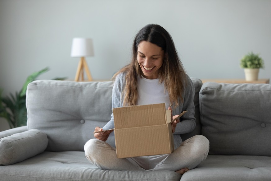 Overjoyed woman seated on sofa hold on lap small cardboard box open parcel client feels satisfied bought goods in internet, happy addressee received package from friend, quick delivery service concept