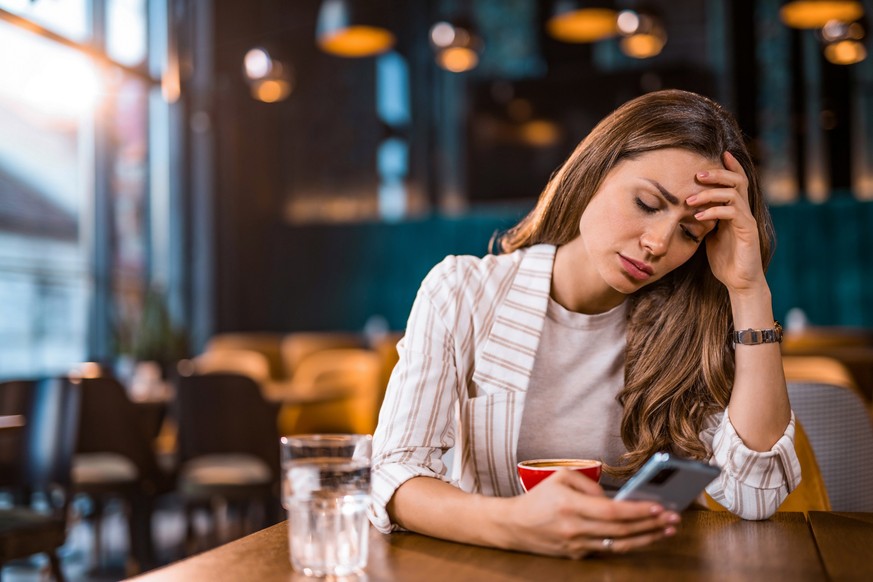 Depressed young woman reading bad news on her phone while sitting at coffee shop.