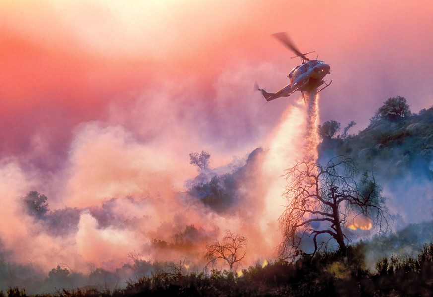A helicopter dropping water on a California wildfire in rugged terrain, backlit by a setting sun filtered through multiple layers of smoke