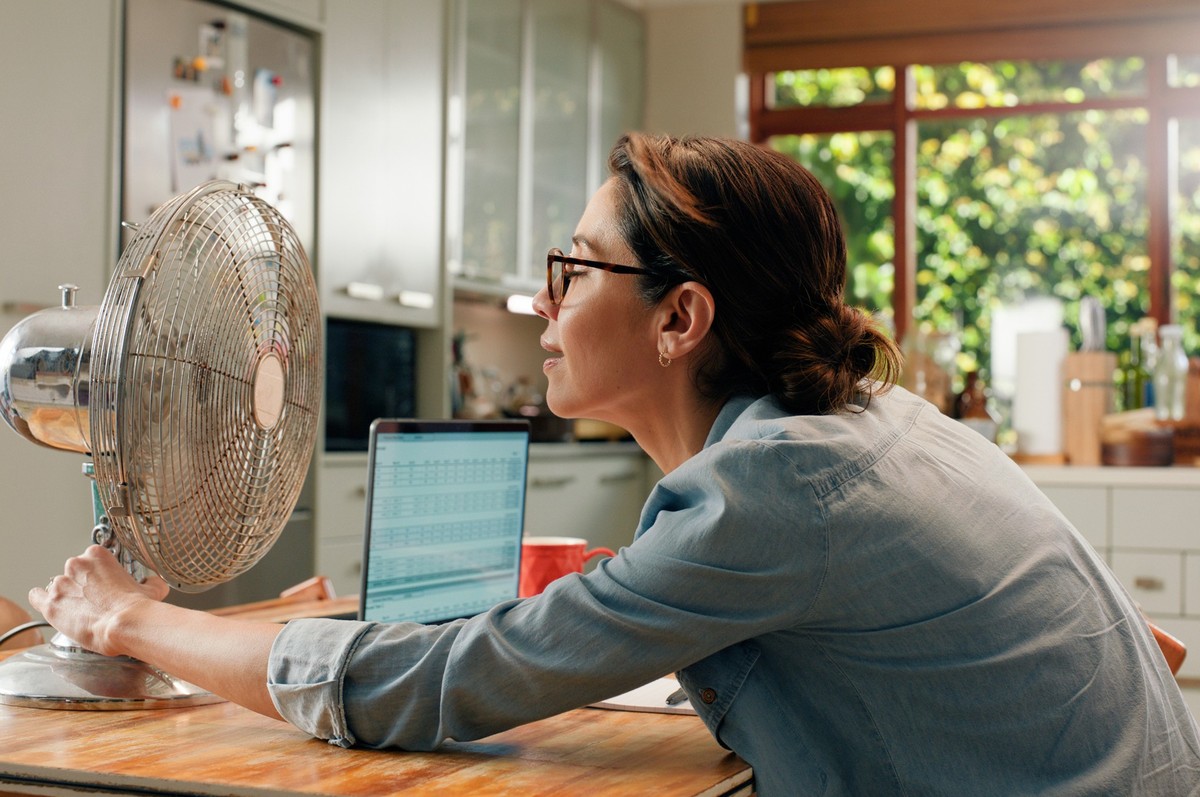 Woman, fan and laptop in home with heat wave as freelance copywriting for remote work, email or kitchen. Female person, over heating and hot weather in apartment with appliance, cool air or summer