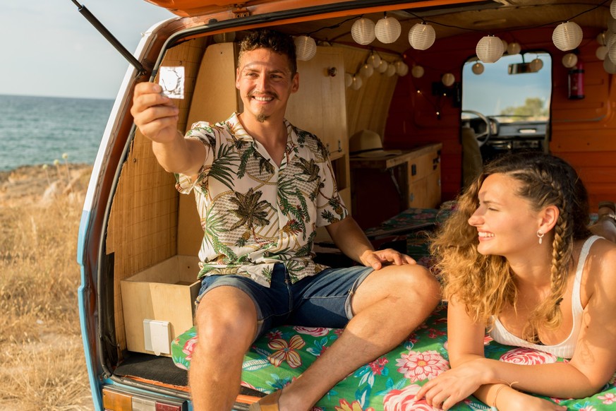 Man holding a condom next to a woman inside a van parked on the beach