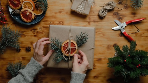 Female hands decorates Christmas gift box with dried orange and Christmas tree branch. Overhead view. Eco friendly Christmas package for holiday gifts.