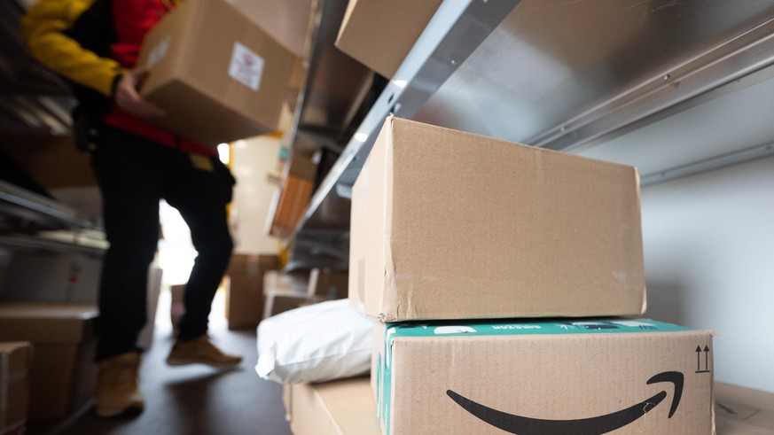 Ein Paket des Online-Händlers Amazon steht in einem DHL-Transporter für die Paketzustellung. Im Hintergrund sortiert ein DHL-Mitarbeiter seine Pakete für die Zustellung.