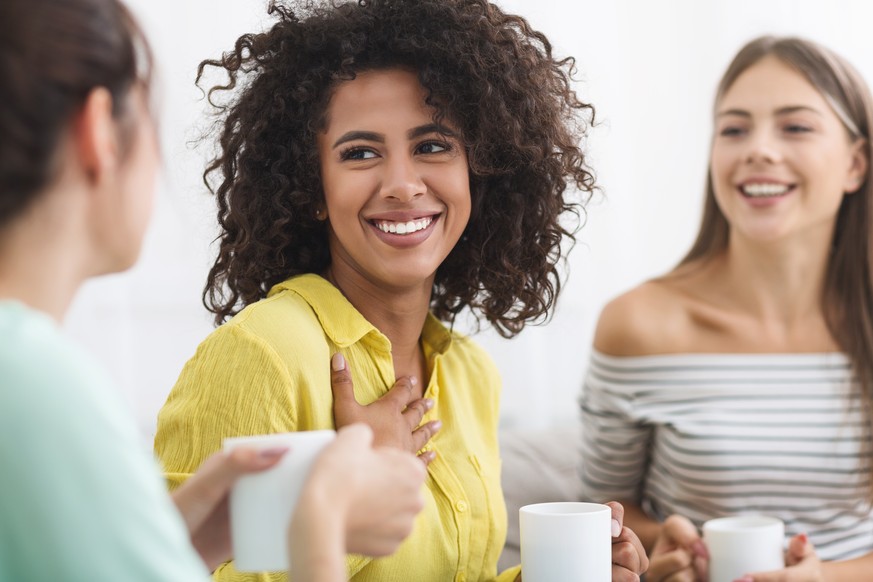 Three happy friends with coffee cups talking about their lifes and relations in living room at home