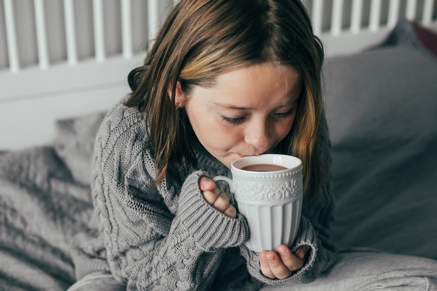 Cozy Winter concept. Child in grey warm sweater drinking hot chocolate while sitting on bed in bedroom. Home lifestyle