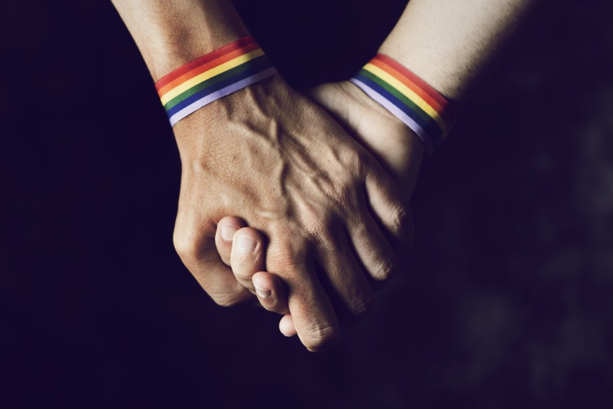 closeup of two caucasian men holding hands with a rainbow-patterned wristban on their wrists