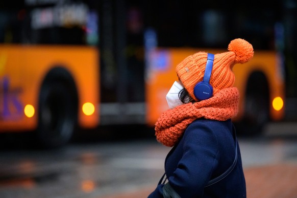 ARCHIV - 06.02.2022, Hamburg: Eine Frau sitzt mit FFP2-Maske, dicker Mütze und Kopfhörern an einem Busbahnhof, während ein Linienbus vorbeifährt. Der dritte Winter mit Sars-CoV-2 steht bevor. Die Ausg ...