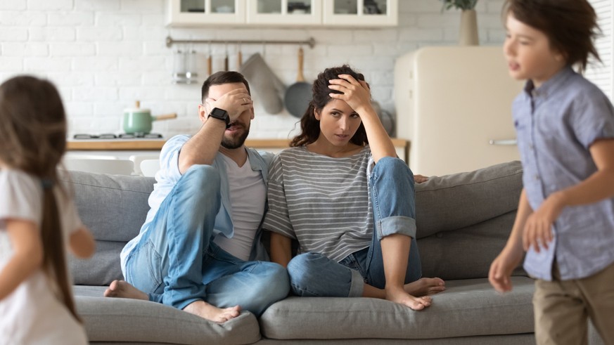 Annoyed young parents sit on couch in kitchen tired from loud two little children running playing, anxious mom and dad relax on sofa at home exhausted from active ill-behaved small preschooler kids