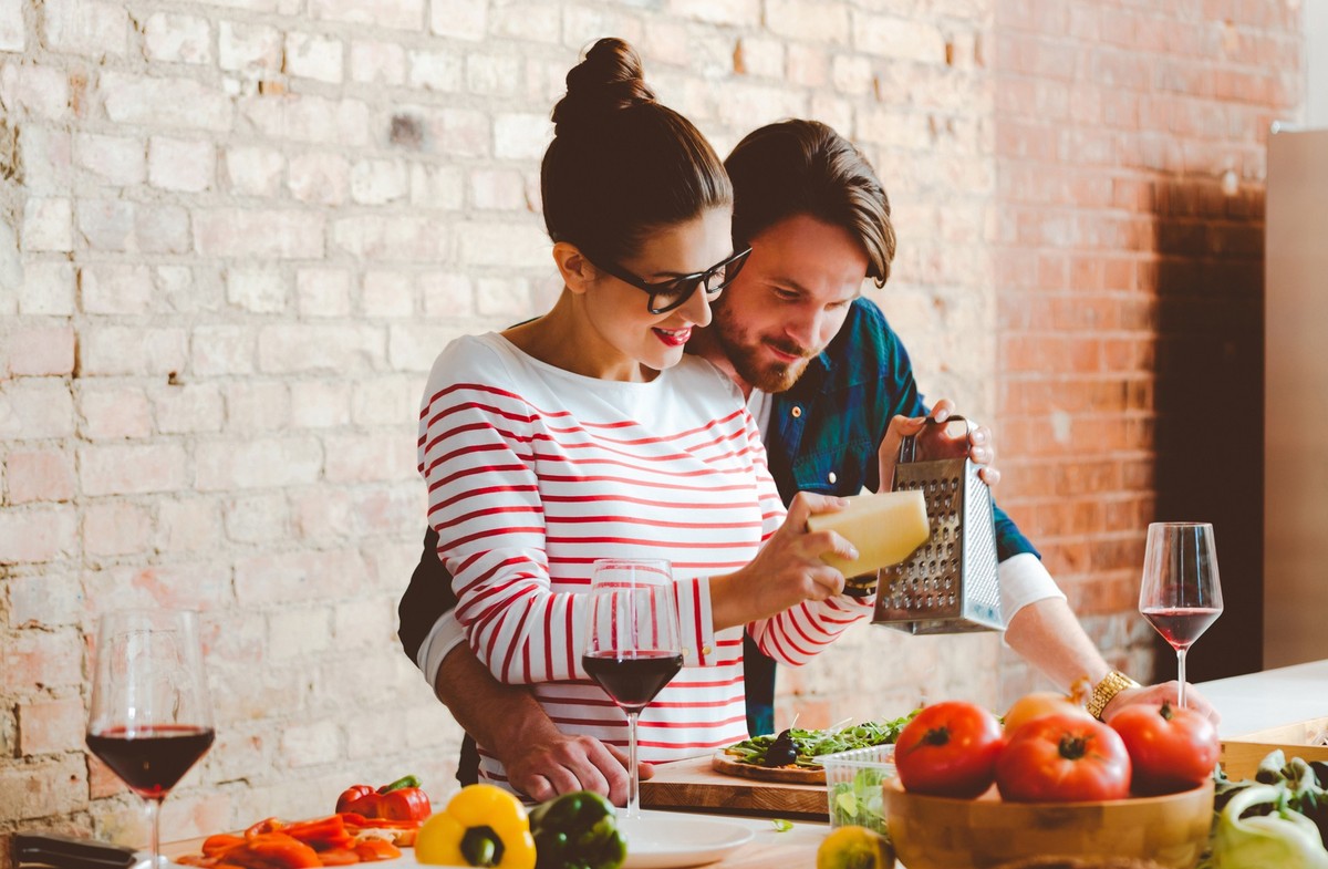 Affectionate couple preparing pizza in the modern kitchen, young woman grating parmesan cheese.
